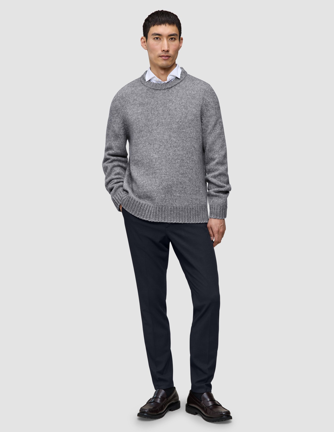 Ein Mann, der einen grauen Pullover mit Rundhalsausschnitt über einem weißen Hemd mit Kragen trägt, dazu eine Motion-Anzughose in der Farbe Slim Midnight Blue und braune Slipper.